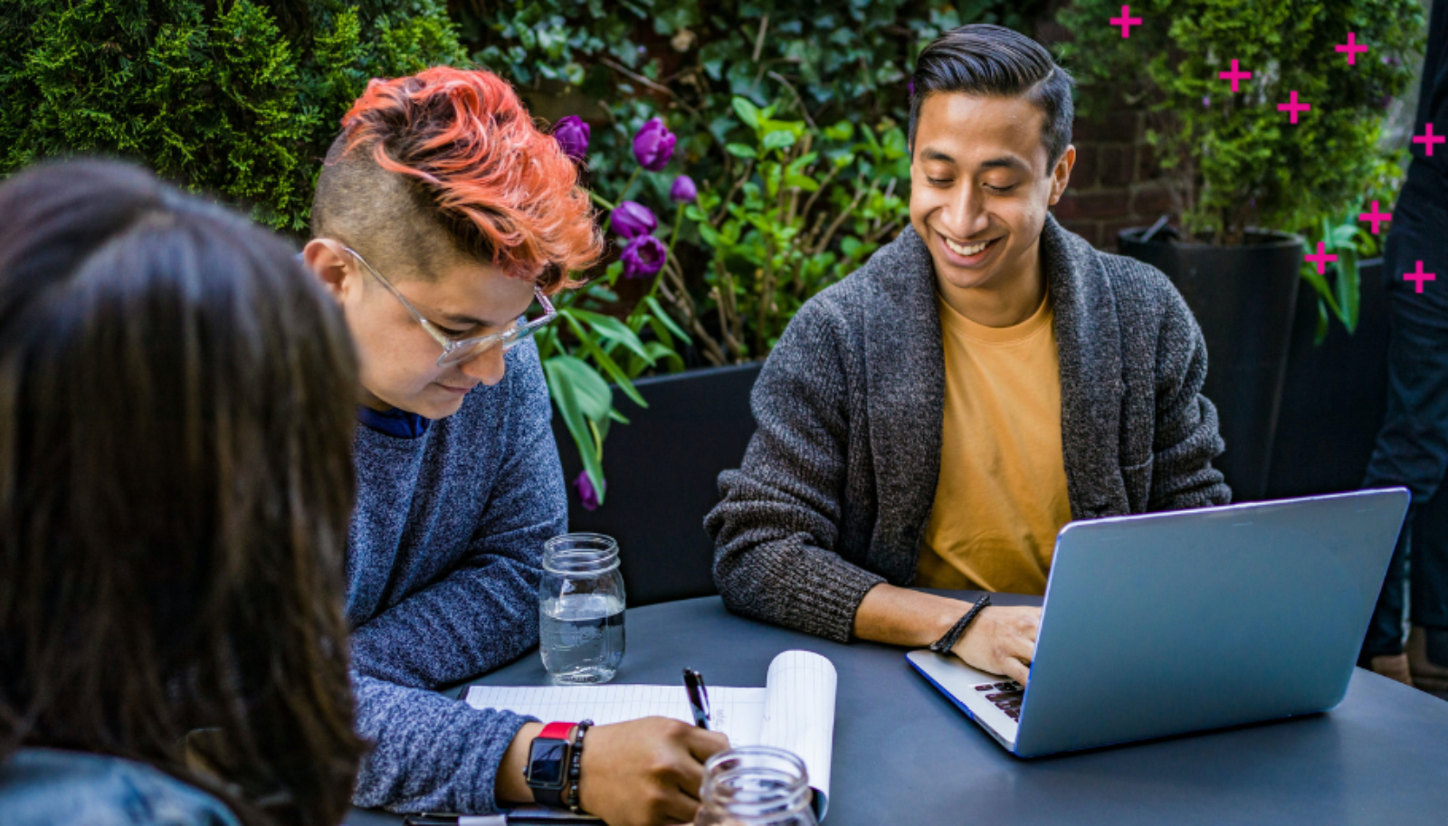 Two people sitting at a table. Left individual is writing on a notepad, the right individual has their hand on a laptop